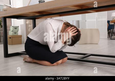 Une jeune femme effrayée se cachant sous le bureau pendant le tremblement de terre Banque D'Images