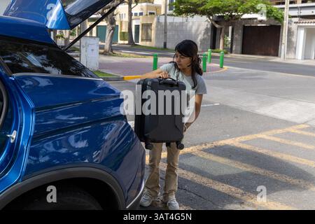 Un adolescent sort une valise d'une voiture bleue. Elle porte une chemise grise. La scène se déroule dans une rue avec quelques arbres en arrière-plan Banque D'Images