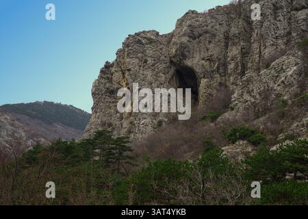 L'image capture un paysage montagneux accidenté avec une falaise rocheuse proéminente et une grande grotte, entourée d'une végétation dense sous un ciel bleu clair Banque D'Images