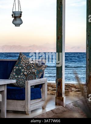 Un coin salon extérieur confortable avec un oreiller décoratif sur une chaise en bois, donnant sur une vue sereine sur l'océan. La scène présente une lanterne suspendue à un p. Banque D'Images