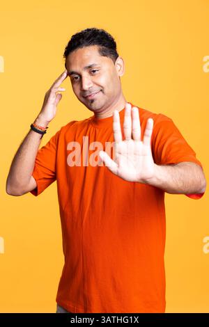 Personne masculine amicale indienne avec un chignon d'homme levant les mains et agitant, saluant ses amis sur fond orange. Jeune adulte dans des vêtements décontractés, joyeux gars satisfait. Banque D'Images