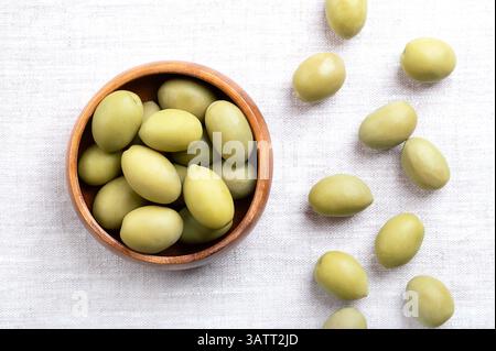 Olives vertes avec fosse, dans un bol en bois sur lin. Olives de table entières, grandes, récoltées avant d'être complètement mûres, avec une saveur plus acidulée et saumeuse. Banque D'Images