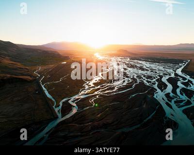 Vue aérienne au coucher du soleil des rivières glaciaires tressées qui s'étendent sur une vaste plaine islandaise, avec des reflets brillants et des silhouettes montagneuses spectaculaires en tressées Banque D'Images