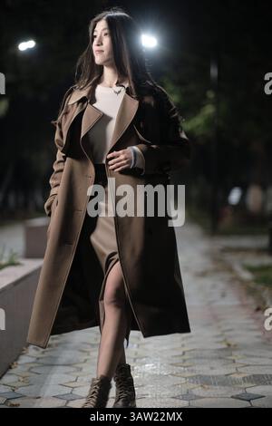 Une charmante jeune femme vêtue d'un long manteau brun et d'un pull léger descend une rue très éclairée la nuit. Banque D'Images