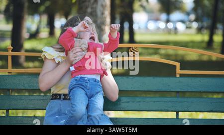 Mère attentionnée réconfortant enfant pleureur sur le banc du parc pendant l'après-midi d'été lumineux Banque D'Images