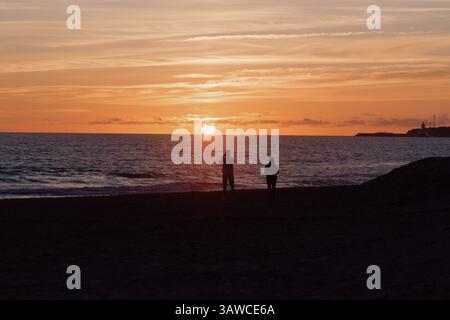 Couple marchant ensemble, silhouetté contre un coucher de soleil orange vibrant, debout pieds nus sur le rivage sablonneux avec des vagues océaniques tranquilles Banque D'Images