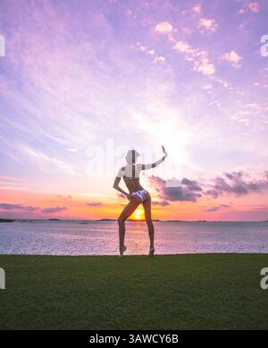 Femme sportive en bikini faisant la pose dansante sur l'herbe devant un beau coucher de soleil sur la mer. Mode de vie actif en voyageant, en poursuivant le lever du soleil. Banque D'Images