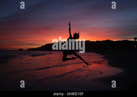 Silhouette de femme sportive en faisant un saut de danse spectaculaire sur la plage en face d'un beau coucher de soleil sur la mer. Mode de vie actif par voyage Banque D'Images