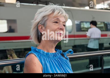 Une femme d'âge mûr vêtue d'une robe bleue dans une gare, attendant un train. Un vrai moment dans le voyage Banque D'Images