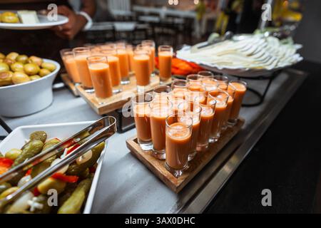 Gros plan d'une table de restauration avec des rangées de petits verres remplis de soupe glacée de gazpacho à l'orange, de cornichons, d'olives vertes et de tranches de fromage disposées dedans Banque D'Images