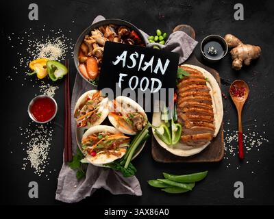 Ensemble de plats chinois sur fond sombre : canard aigre-doux, soupe de nouilles, petits pains cuits à la vapeur. Banquet ou buffet asiatique, vue de dessus. Banque D'Images
