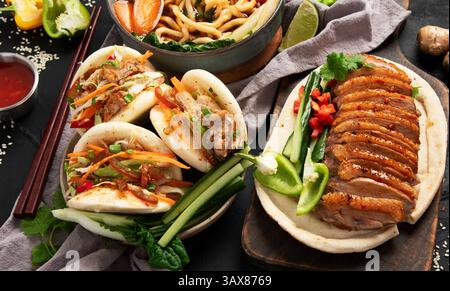 Ensemble de plats chinois sur fond sombre : canard aigre-doux, soupe de nouilles, petits pains cuits à la vapeur. Banquet ou buffet asiatique, vue de dessus. Banque D'Images