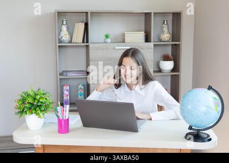 Jeune femme travaillant à partir du bureau à domicile avec ordinateur portable. Une jeune femme travaille à la maison, assise à son bureau avec un ordinateur portable. Elle sourit et tient Banque D'Images
