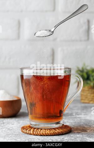 Sucre blanc tombant de la cuillère à café dans une tasse en verre de thé chaud placé sur des montagnes russes en osier contre le mur de briques comme fond Banque D'Images