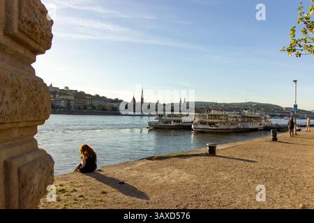 Vue panoramique sur la rivière du Danube à Budapest avec bateaux de croisière amarrés, paysage urbain historique en arrière-plan, et les gens se relaxant sur la promenade Banque D'Images