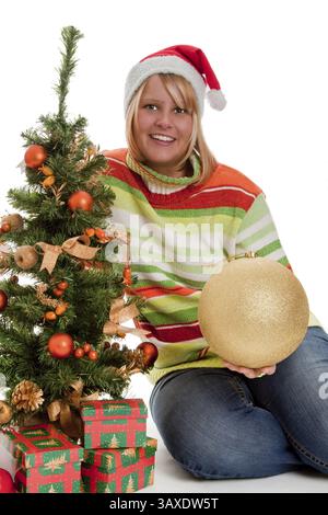 Jeune femme dans le chapeau de Père Noël assis devant un arbre de Noël décoré avec des cadeaux - coupés Banque D'Images