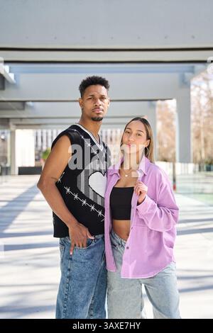 Jeune couple multi ethnique à la mode posant ensemble sous une structure architecturale moderne dans un cadre urbain Banque D'Images