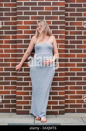Belle femme enceinte posant contre un mur de briques tout en touchant son ventre à la lumière naturelle Banque D'Images