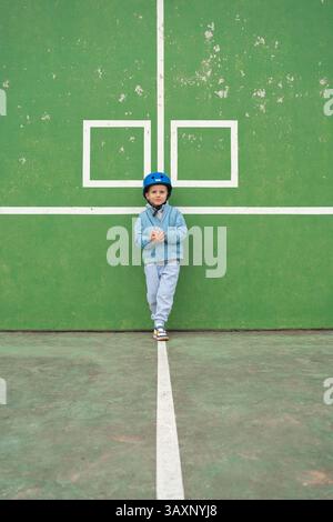 Enfant cool posant sur une aire de jeux, portant un casque bleu et une tenue décontractée. Le mur vert avec des lignes de cour ajoute un fond géométrique moderne. Parfait pour Banque D'Images