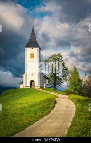 Paysage de printemps et église alpine sur la colline. Endroit merveilleux avec route de gravier rurale et église Saint Primoz en arrière-plan, village de Jamnik, Slovénie Banque D'Images