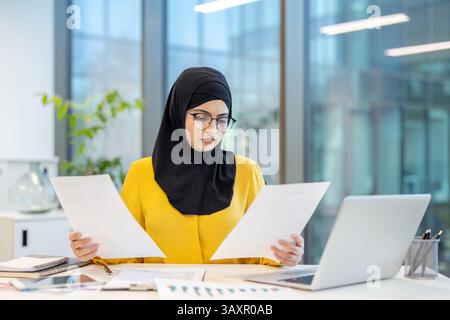 Une femme arabe avec des lunettes portant un hijab examine des documents dans un cadre de bureau moderne. Elle semble concentrée et concentrée au travail. Banque D'Images