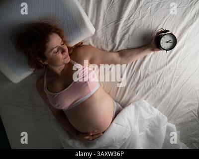 Une femme enceinte se couche dans son lit et tient un réveil. Banque D'Images