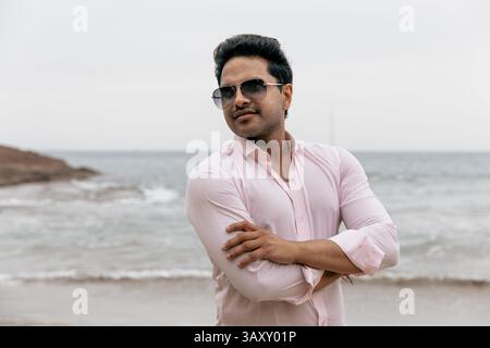 Homme confiant en lunettes de soleil avec bras pliés debout par l'océan. Banque D'Images