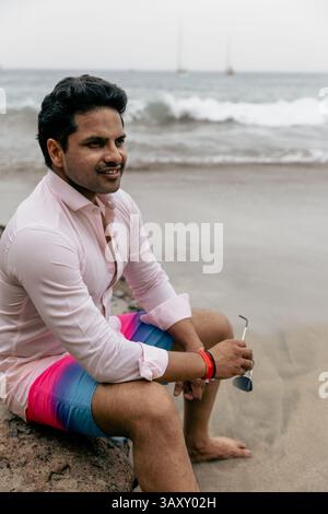 Un homme est assis sur un rocher sur une plage portant une chemise rose et un short bleu et rose. Il tient ses lunettes de soleil Banque D'Images