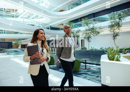 Deux collègues professionnels traversent un espace de cabinet d'avocats moderne avec une lumière naturelle lumineuse et un design contemporain. Ils sont engagés dans la conversation, Banque D'Images