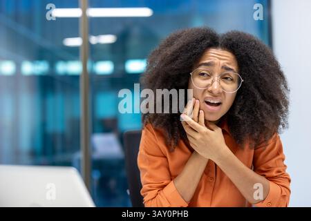 Une jeune femme avec des lunettes est visiblement dans la douleur, serrant sa mâchoire comme si elle éprouvait un mal de dents. Le cadre suggère un environnement de bureau, avec un arrière-plan flou. Banque D'Images