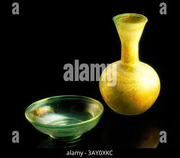Un élégant récipient translucide avec un col gracieux est accompagné d'un plat rond peu profond. Les deux pièces présentent une qualité lumineuse, présentant des textures complexes et une lueur douce sur un fond sombre., Roman, bouteille, 4ème siècle AD, verre, h 9 7/8 Diam 6 1/2, Art classique avec une touche moderne réinventée par Artotop Banque D'Images