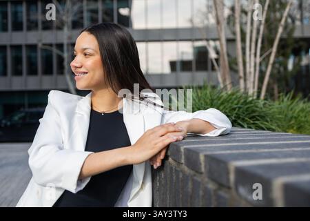 Une femme hispanique souriante dans un blazer blanc profite d'une pause à l'extérieur d'un immeuble de bureaux moderne, rayonnant de confiance et de succès dans sa tenue d'affaires. Banque D'Images
