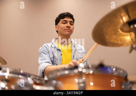Un batteur portant une veste en denim et une chemise jaune joue habilement de la batterie dans un studio d'enregistrement bien éclairé, capturant l'essence de la création musicale Banque D'Images
