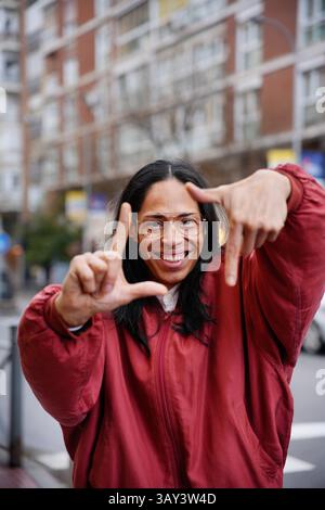 Un homme joyeux dans une veste rouge crée un cadre avec leurs mains, mettant l'accent sur la concentration et la perspective. L'arrière-plan urbain flou rehausse l'animation, bu Banque D'Images