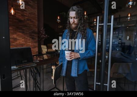 Un homme aux cheveux longs tient une planche à roulettes et une tasse de café à emporter tout en se tenant debout dans un café élégant. L'environnement dispose d'un éclairage moderne et rusti Banque D'Images