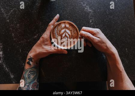 Une vue de dessus de mains tatouées tenant une tasse de latte avec art en mousse complexe sur une table sombre. La scène reflète un mélange de créativité et de relaxation Banque D'Images