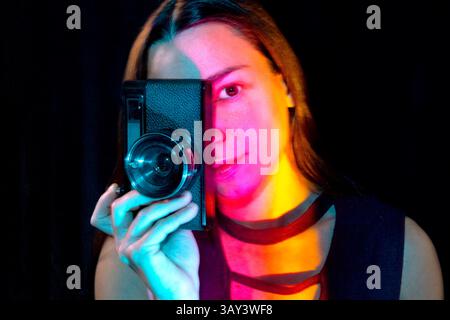 Une femme tient un appareil photo vintage, tandis que des lumières colorées vibrantes illuminent son visage. Le contraste et les ombres créent une atmosphère artistique et mystérieuse. Banque D'Images
