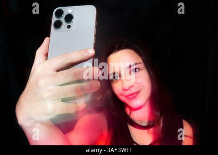 Une femme souriante en prenant un selfie avec un smartphone sur fond noir. La vue rapprochée met en évidence le téléphone et son expression agréable Banque D'Images