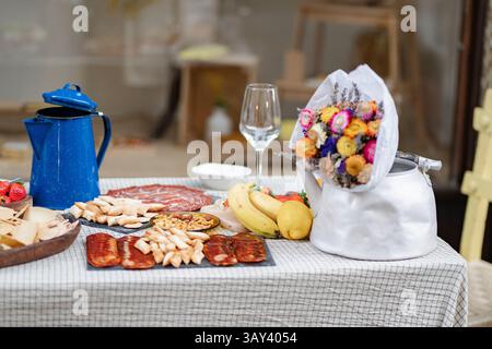 Un pique-nique rustique sur une nappe à carreaux, avec des fleurs séchées dans une bouilloire, des fruits vibrants, des fromages assortis, des charcuteries et un café bleu Banque D'Images