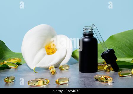 Mat bouteille noire de sérum cosmétique à côté d'un lys blanc calla, avec une pipette et des capsules oméga de suppléments dorés dispersés sur un blu doux Banque D'Images