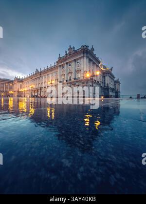 Majestueux Palais Royal de Madrid avec façade illuminée au crépuscule. L'architecture est reflétée dans une surface imbibée de pluie, créant un reflet serein Banque D'Images
