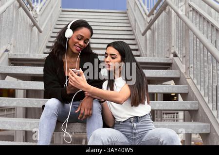 Deux amis s'assoient sur les escaliers extérieurs partageant de la musique avec des écouteurs. L'un sourit tandis que l'autre montre un smartphone, créant un atmos joyeux et détendu Banque D'Images