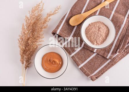 Vue de dessus de poudres d'argile cosmétique dans des bols à côté d'une plante ornementale, présenté avec une cuillère en bois sur un tissu texturé, idéal pour les soins du visage Banque D'Images