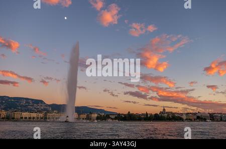 Magnifique coucher de soleil illuminant les nuages sur l'emblématique Jet d'eau de Genève, avec le lac Léman au premier plan et la ligne d'horizon de la ville derrière. Banque D'Images