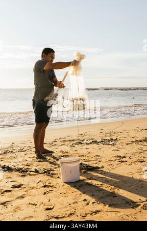 Un pêcheur se tient sur une plage de sable sous un ciel dégagé, triant ses prises d'un filet. Le soleil projette de longues ombres, et l'océan offre un arrière-plan serein Banque D'Images