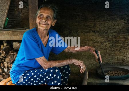 Une femme âgée dans une chemise bleue et un pantalon à pois torréfie des grains de café balinais dans un cadre rustique. Son sourire serein reflète la sagesse et le simpl Banque D'Images