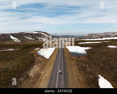 Une route droite traverse les hautes terres islandaises, avec des taches de neige et une voiture isolée soulignant la vaste nature intacte sous un ciel bleu. Banque D'Images