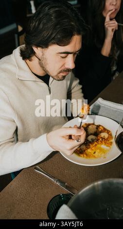 Un homme mange une assiette de nourriture avec une fourchette. Il porte une chemise blanche et est assis à une table Banque D'Images