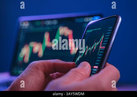 Gros plan des mains tenant le smartphone affichant un graphique de bougies boursières, avec l'écran d'ordinateur portable en arrière-plan. Trader analysant les graphiques financiers sur un téléphone tard dans la nuit Banque D'Images