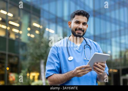 Un sympathique travailleur de la santé dans les gommages bleus tient une tablette tout en souriant, debout devant un bâtiment moderne, peut-être un établissement médical. Banque D'Images